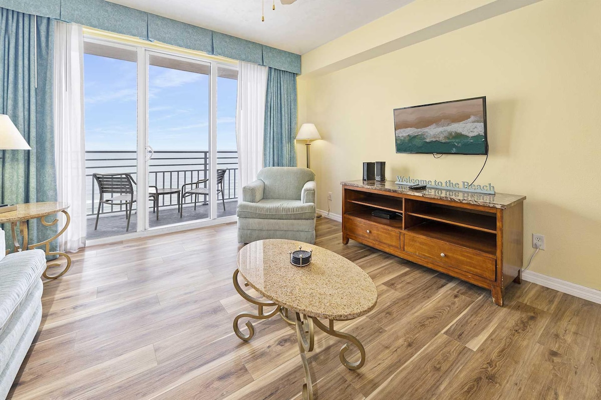 A welcoming living area features a comfortable armchair and a stylish coffee table positioned on light wood flooring. A television is mounted on the wall, and sliding glass doors lead to an oceanfront balcony, offering expansive views of the beach and water.