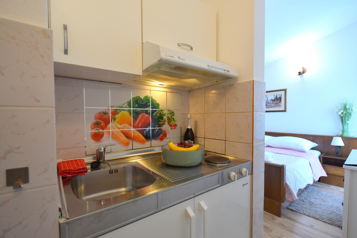 The kitchenette features a compact layout with a stainless steel sink and stove. A bowl of fresh fruit is placed on the counter, alongside a decorative backdrop of colorful vegetables. A cozy sleeping area is partially visible in the background, enhancing the apartment's functionality.