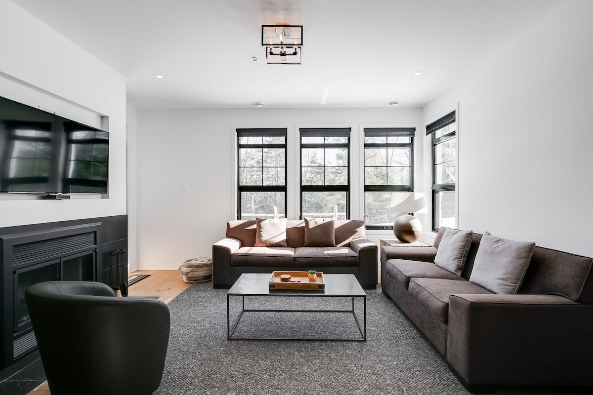 A modern living area features a comfortable sectional sofa accompanied by a sleek armchair. Natural light enters through multiple windows, illuminating the space. A low coffee table resides on a textured rug, with a flat-screen television mounted on the wall above a cozy fireplace.