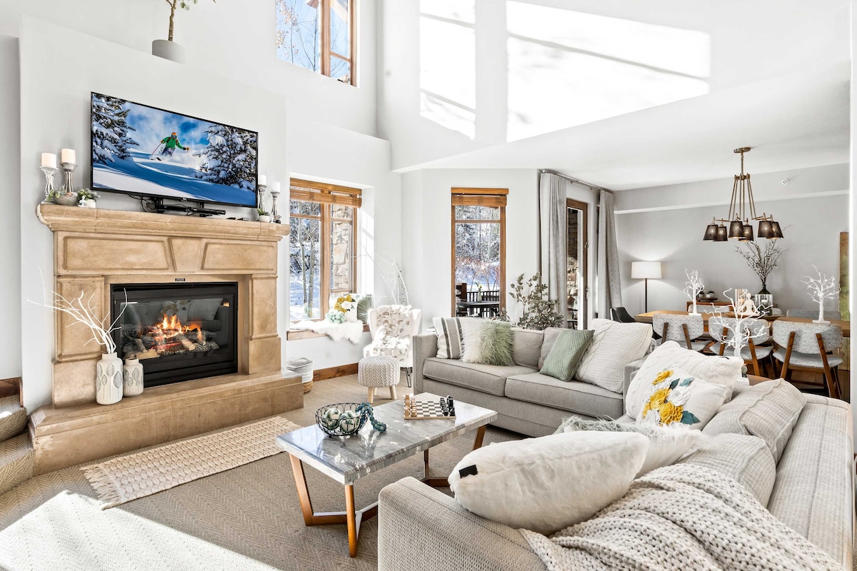 A welcoming living area features a stone fireplace with a mounted television above. Cozy seating arrangements with light grey sofas surround a glass coffee table. Large windows allow natural light to fill the space, while a dining area with a wooden table and upholstered chairs is visible in the background.