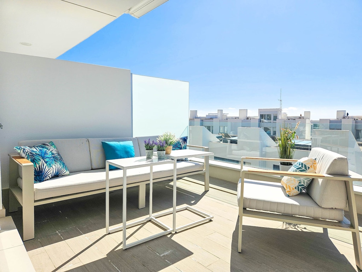 A spacious terrace is furnished with modern outdoor seating, including a sofa and a table. Potted plants are arranged on the surface, and decorative pillows in vibrant colors add a touch of style. Clear skies and surrounding rooftops are visible in the background.