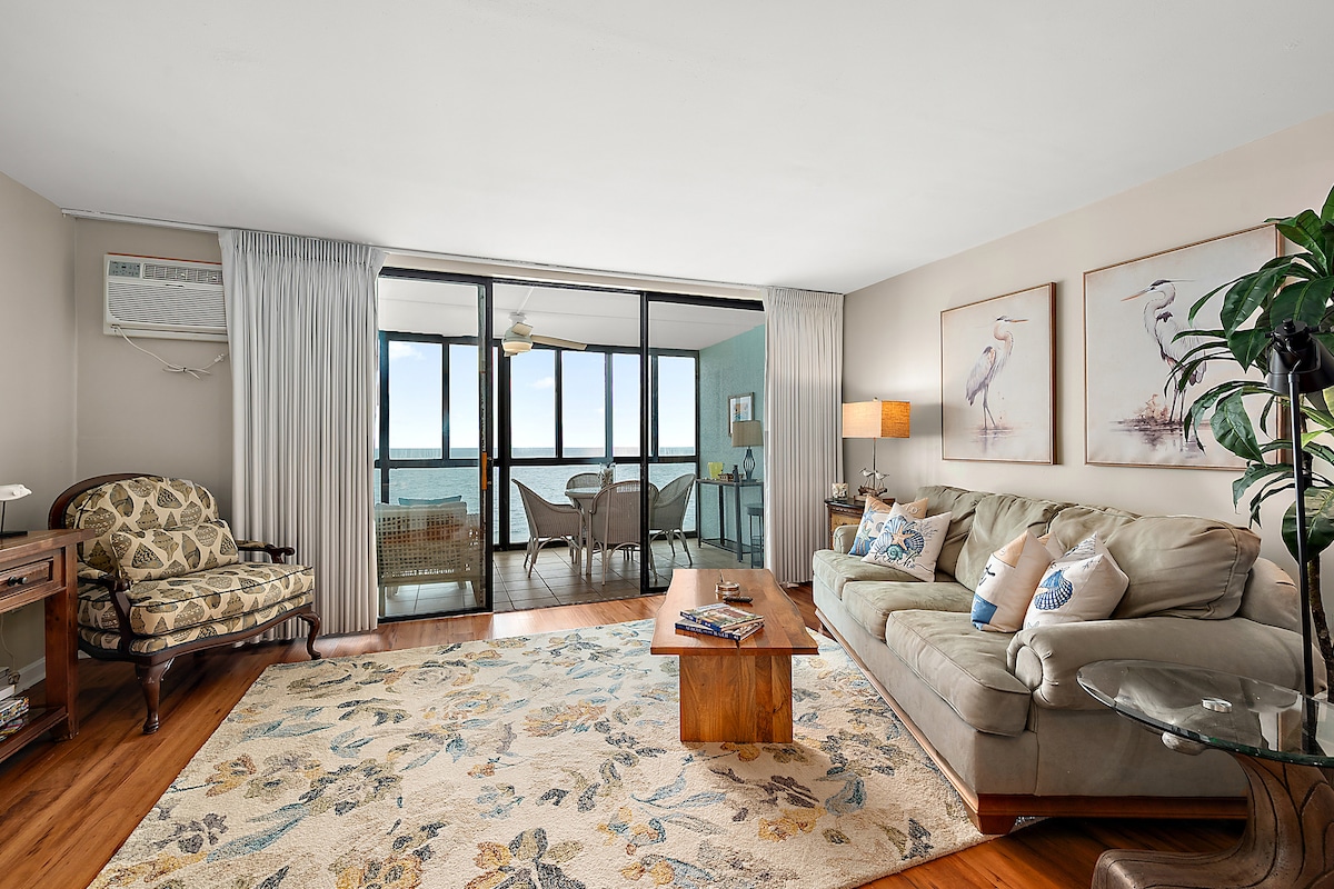 A living area features a spacious sofa and accent chairs arranged invitingly. Natural light filters in through large sliding glass doors, offering views of the ocean. Decorative artwork adorns the walls, and a light-colored area rug adds a touch of warmth to the hardwood floor.