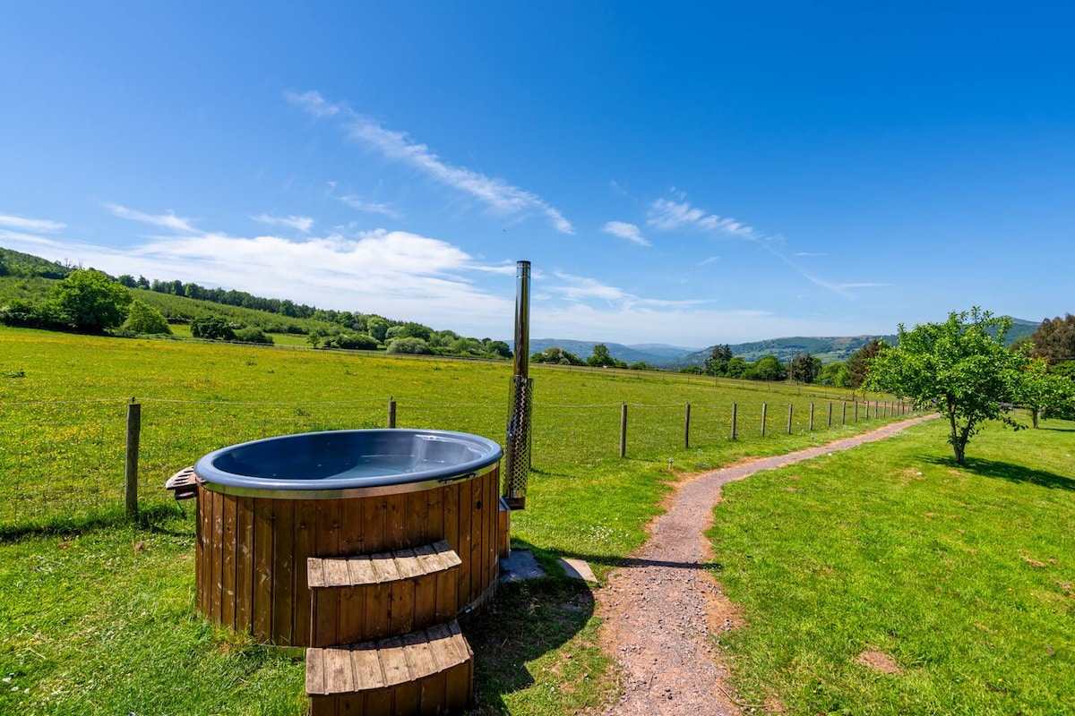 A wooden hot tub is positioned on a grassy area, showcasing a rustic design. Steps lead up to the hot tub, surrounded by a lush green landscape. Fields extend into the distance, beneath a clear blue sky, framed by distant hills.