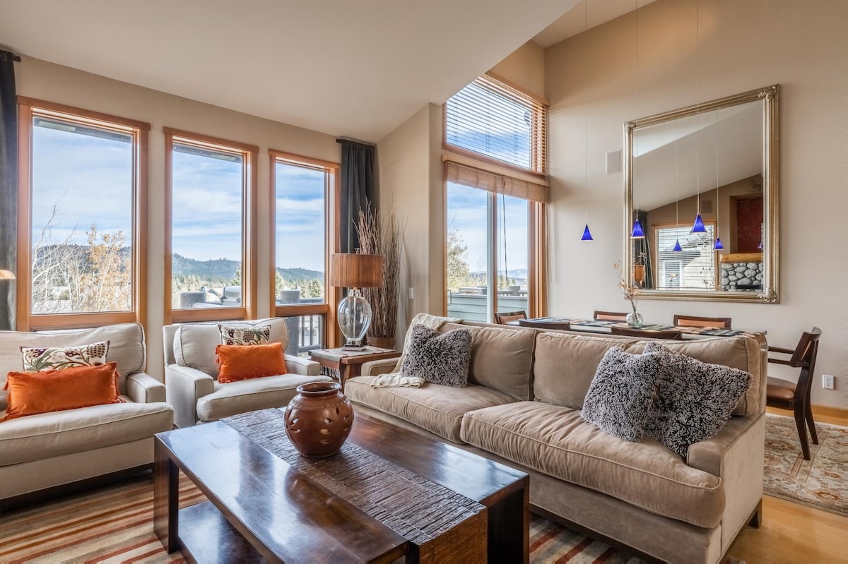 A spacious great room features a comfortable sofa adorned with textured throw pillows. Large windows provide natural light and showcase views of the surrounding landscape. A stylish mirror reflects the ambiance, while a wooden coffee table sits at the center, complemented by decorative accents.