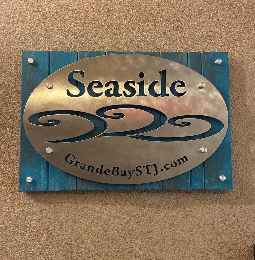 A wall-mounted sign displaying the word 'Seaside' along with an emblem of stylized waves is presented. The sign features a metallic finish with a turquoise background and is secured with decorative screws.