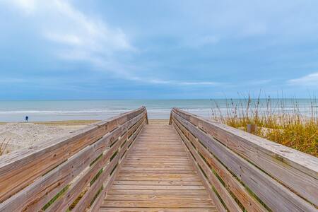 A wooden boardwalk extends toward the ocean, bordered by tall grasses on either side. The calm sea is visible in the distance under an overcast sky, with hints of blue peeking through the clouds. A solitary figure can be seen walking along the beach to the left.
