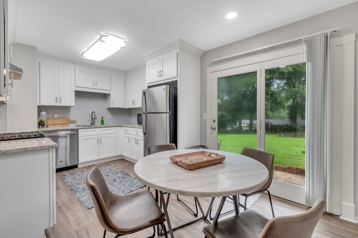 A bright kitchen features white cabinetry, granite countertops, and stainless steel appliances. A round table with four chairs centers the space, while a sliding glass door offers views of the green outdoor area. Natural light enhances the open and airy atmosphere.