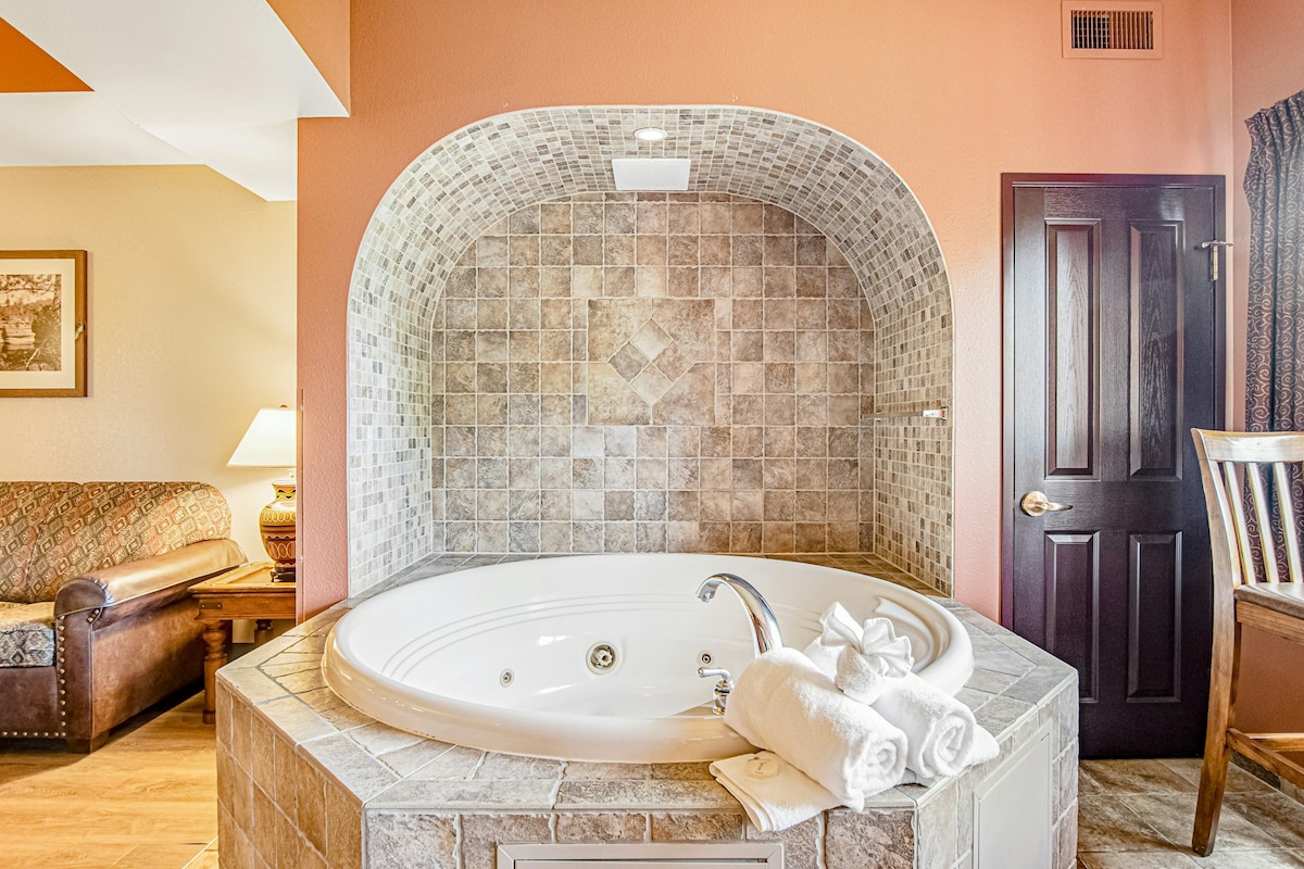 A jetted hot tub is set against a textured stone backdrop, providing a relaxing focal point in the room. Soft, white towels are neatly arranged beside the tub. The space is complemented by warm orange walls and a cozy sitting area visible in the background.