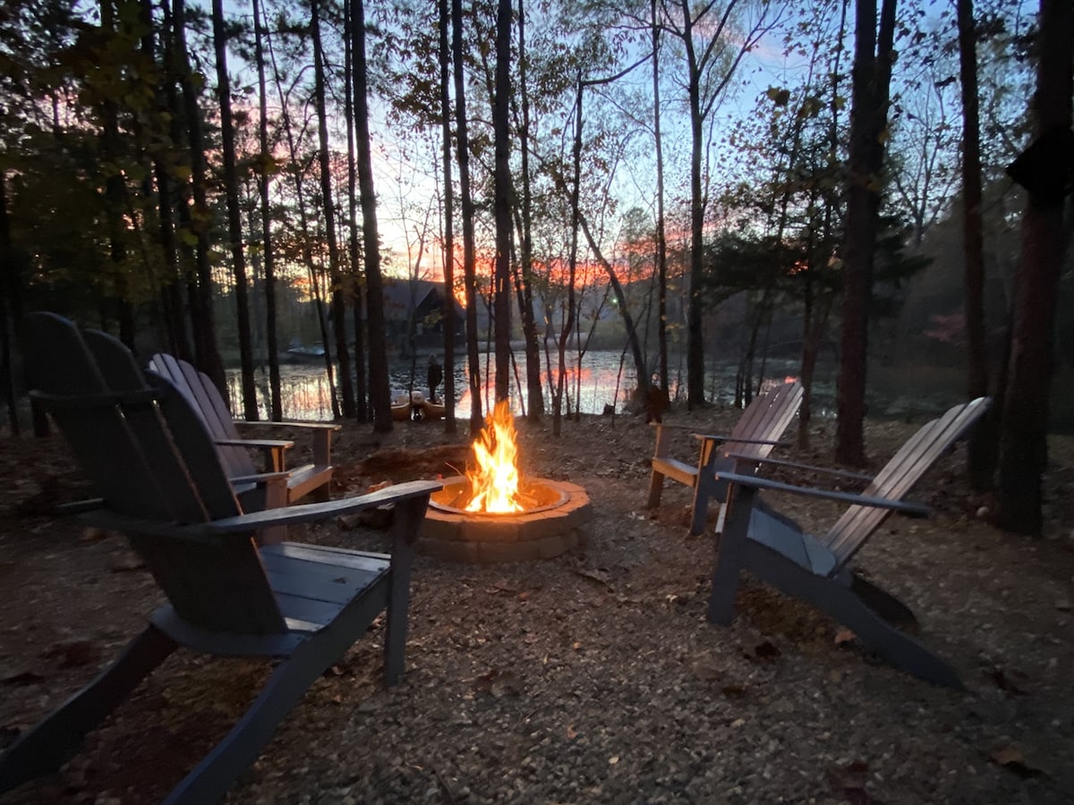 A fire pit surrounded by four wooden Adirondack chairs is illuminated by a gentle fire. The scene features trees in the background, framing the serene pond and distant landscape under a colorful sunset sky.
