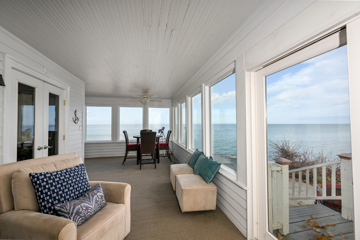 A sunlit sunroom features large panoramic windows offering views of Lake Erie. A comfortable sofa with decorative pillows is positioned alongside a small ottoman. A dining set with four chairs is visible, creating a space for meals or relaxation by the water.