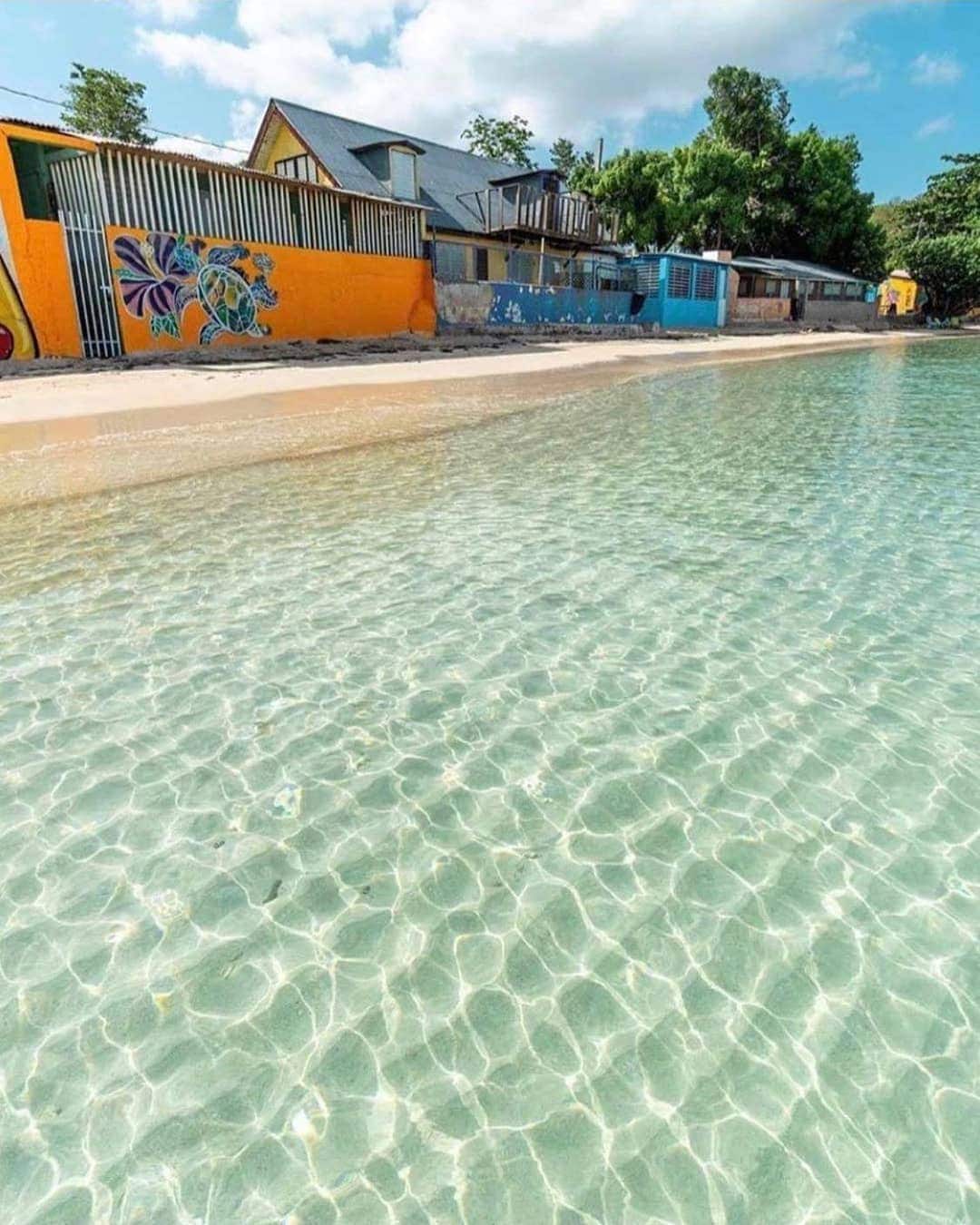 Crystal clear waters stretch along a sandy shoreline, revealing vibrant reflections of the surrounding landscape. A colorful mural decorates a nearby building, adding a pop of color to the serene seaside environment. The area is enhanced by the gentle waves lapping at the beach.