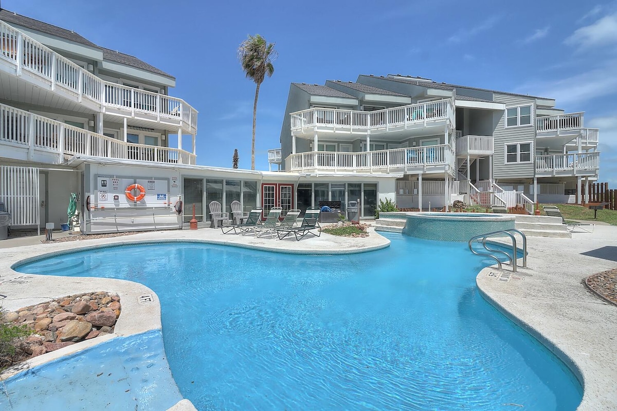 A curved swimming pool is surrounded by lounge chairs located on a well-maintained pool deck. The building features multiple levels with balconies that overlook the pool area. Palm trees can be seen in the background, contributing to the vacation atmosphere.