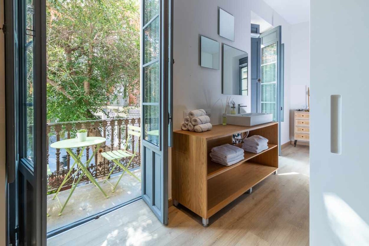 A bright bathroom area is visible, featuring a modern sink and a wooden shelf hosting neatly stacked towels. Two large windows with blue frames open to a small balcony with a bistro table and chairs, allowing natural light to enhance the space.