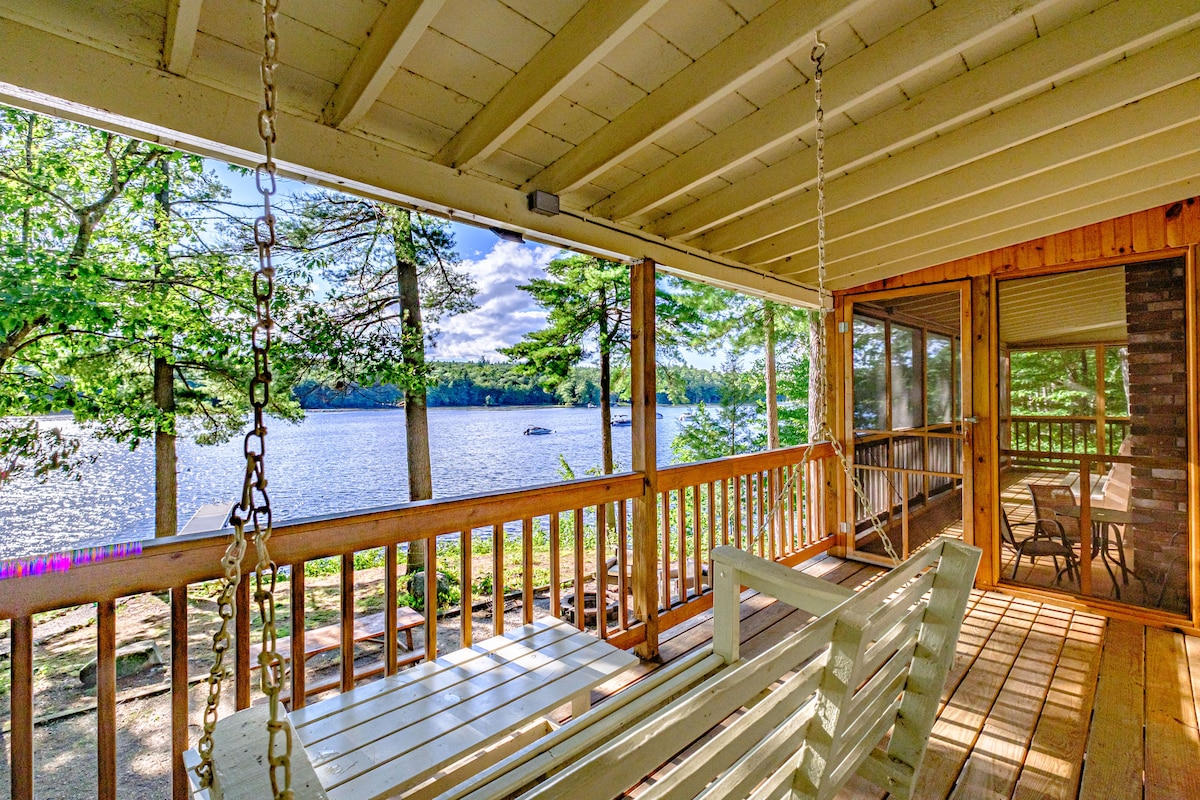 A screened-in porch features a porch swing and bistro set, offering views of the lake and surrounding trees. The wooden decking provides a natural look while inviting relaxation, with sunlight filtering through the trees.
