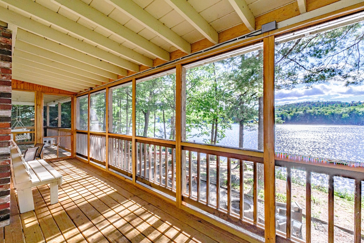 A screened-in porch features wooden flooring and ample seating, overlooking sparkling waters and lush trees. Sunlight streams through the screens, creating a warm, inviting atmosphere. This space provides a serene view of the lake, making it ideal for relaxing and enjoying nature.