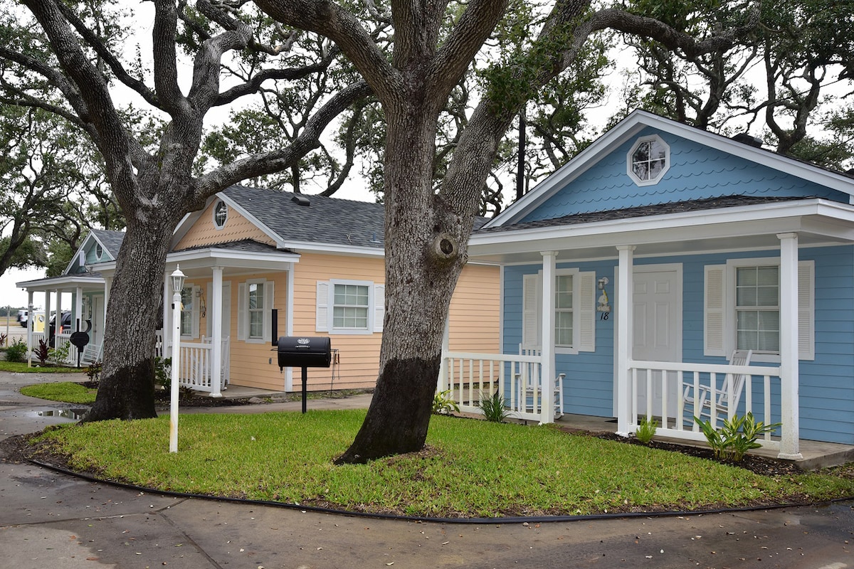 A row of colorful cottages is positioned along a circular pathway, surrounded by lush green grass and mature oak trees. Each cottage features a covered porch, with prominent windows allowing ample natural light. Landscaping adds a touch of greenery to the inviting outdoor area.