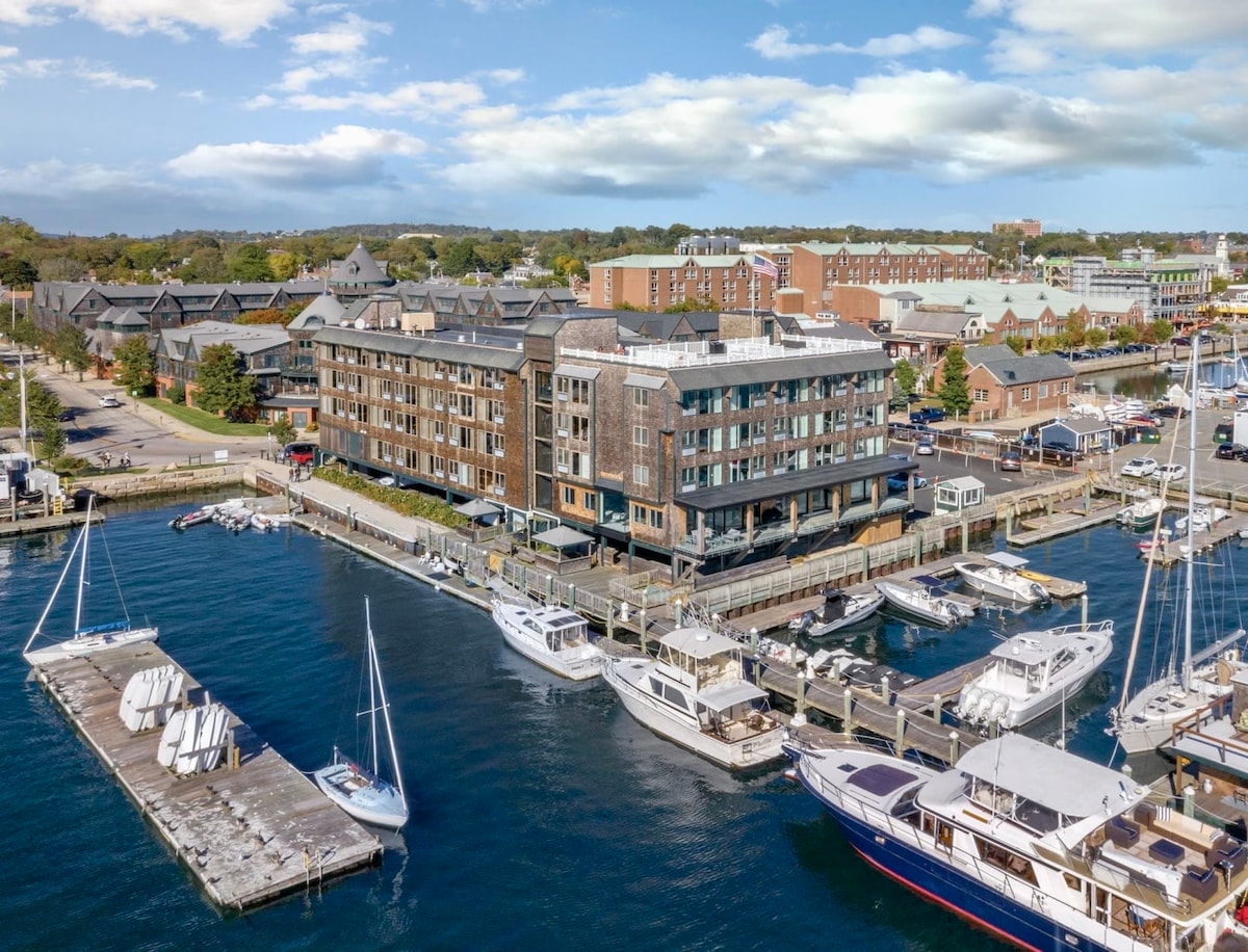 The image shows an aerial view of a waterfront hotel positioned alongside a marina. Various boats are docked nearby, and the surrounding area features a mix of buildings and greenery. The sky is partly cloudy, enhancing the bright atmosphere of the scene.