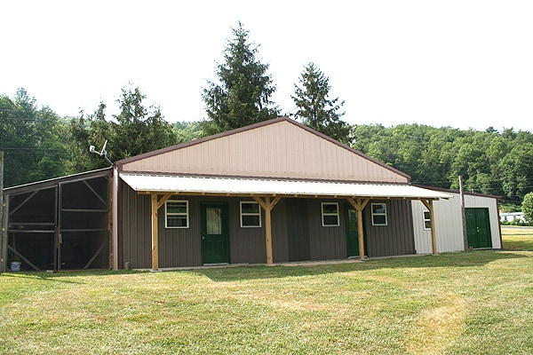A spacious building is shown, featuring a combination of gray and tan siding with a metal roof. Green doors frame the structure, which is flanked by trees and open grass areas, suggesting a connection to nature.