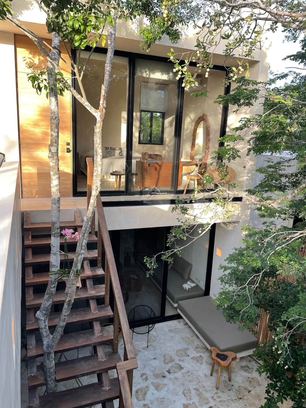 An outdoor view showcases the villa's multi-level structure surrounded by trees. A wooden staircase leads to a private terrace with comfortable seating. Large glass doors create a seamless connection to the interior, allowing natural light to fill the space.