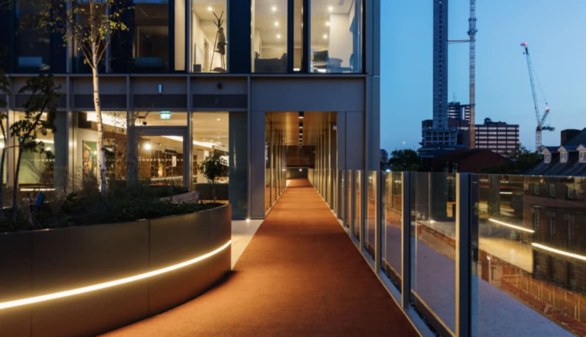 An illuminated pathway leads through a modern building towards a serene outdoor area. Soft lighting highlights the elegant curves of the walkway, surrounded by greenery and glass railings. The evening sky adds a calm ambiance, contrasted with city structures in the background.