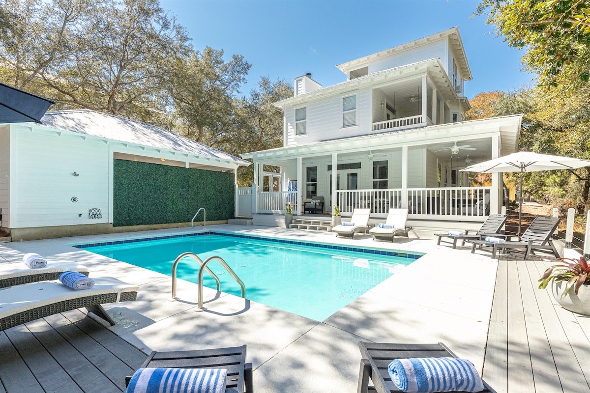 The image shows a private heated pool surrounded by a spacious wooden deck. Lounge chairs are neatly arranged around the pool, and a sun umbrella provides shade. The home’s white exterior is visible in the background, framed by greenery and trees.