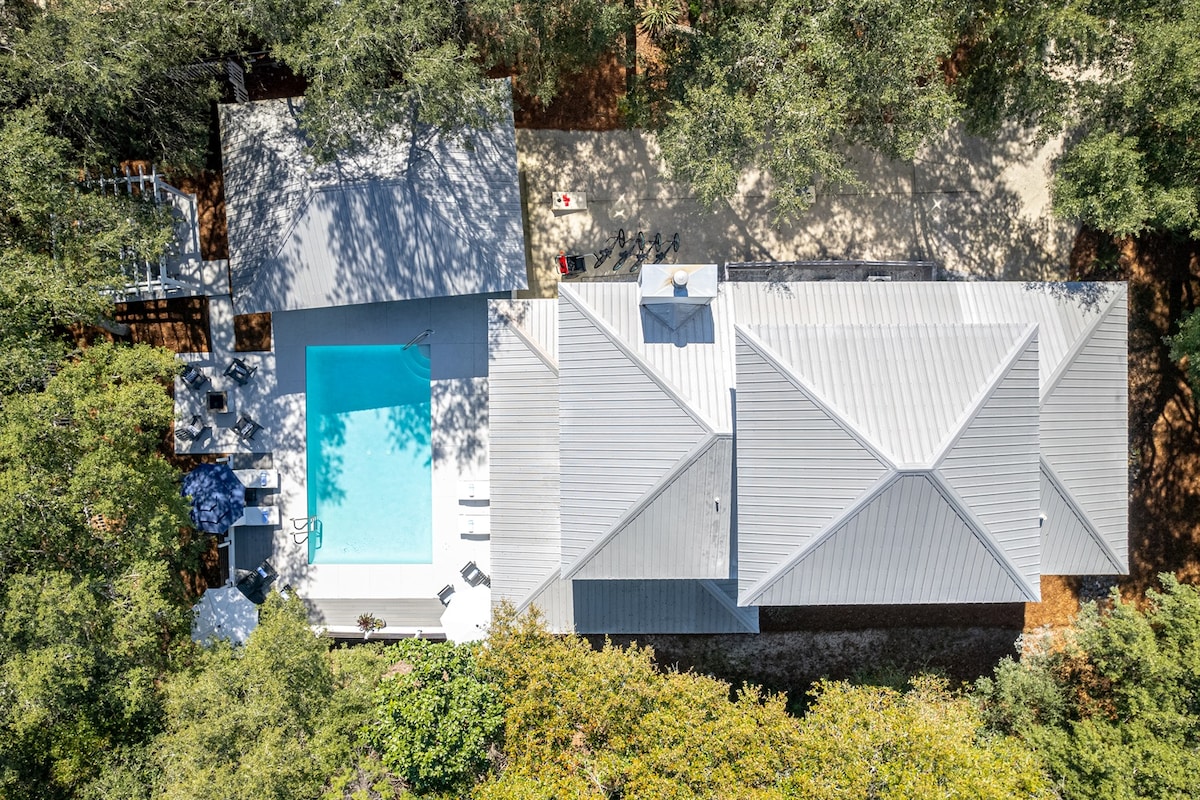An aerial view of a modern home surrounded by trees, featuring a private heated pool and spacious patio. The outdoor space includes loungers and an umbrella. The rooflines of the house showcase a contemporary design, blending harmoniously with the natural landscape.