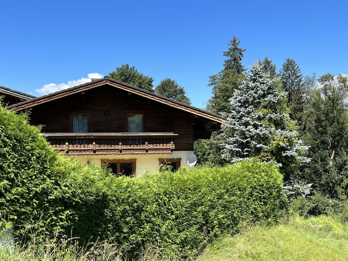 The exterior of the house displays wooden architecture, blending into the surrounding greenery. A balcony is visible on the upper level, overlooking lush trees. The sky above is clear and blue, creating a serene backdrop for the property.