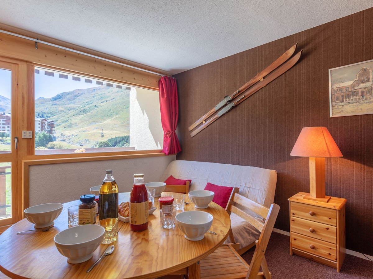 A dining area is shown with a wooden table set for breakfast, featuring bowls and various condiments. A cozy couch is positioned against the wall, with two wooden skis displayed above. Natural light enters through the window, offering a view of the mountains.
