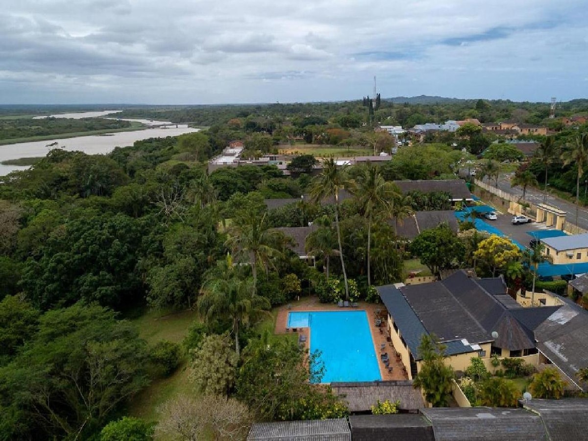 An aerial view showcases a sprawling complex surrounded by lush greenery, featuring a large communal pool with clear blue water. The St Lucia Lake can be seen in the background, alongside a scenic landscape dotted with trees and buildings.