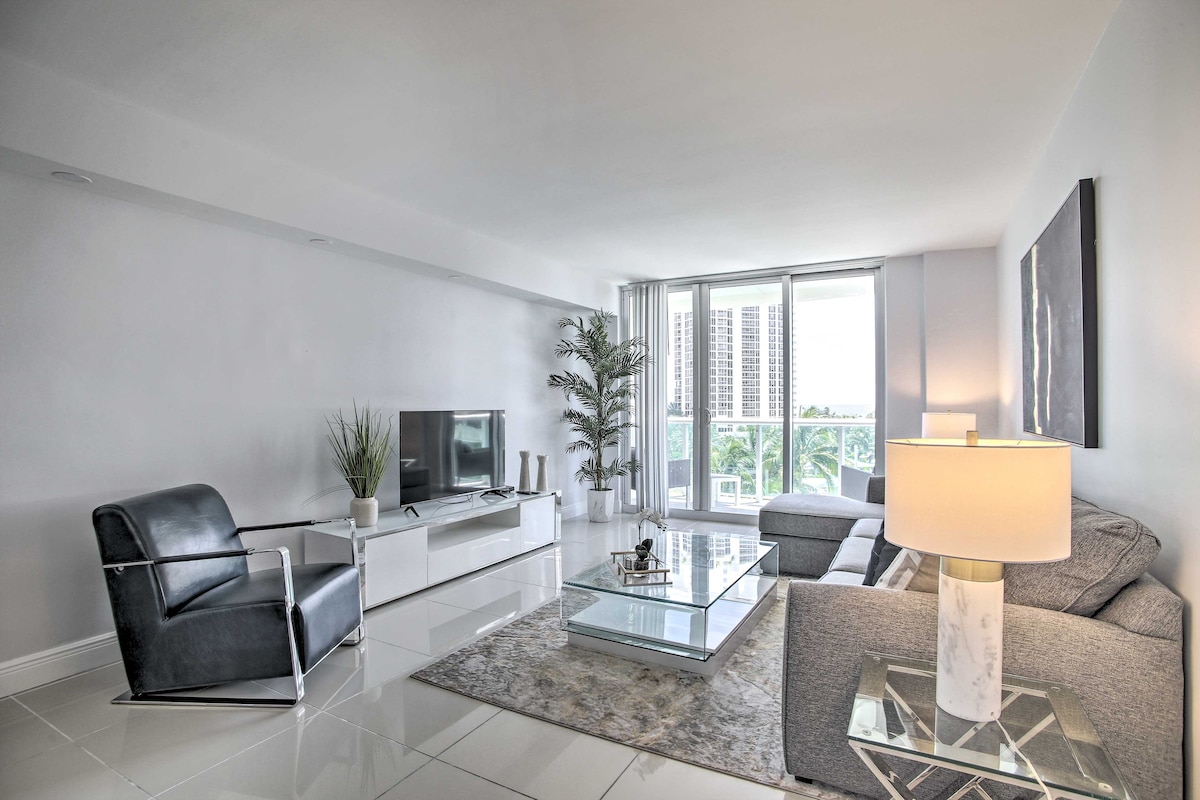 A spacious living area is highlighted by large windows, allowing natural light to fill the room. A comfortable gray sofa faces a modern coffee table and entertainment unit, while a sleek black armchair adds a touch of elegance. A decorative plant enhances the ambiance.