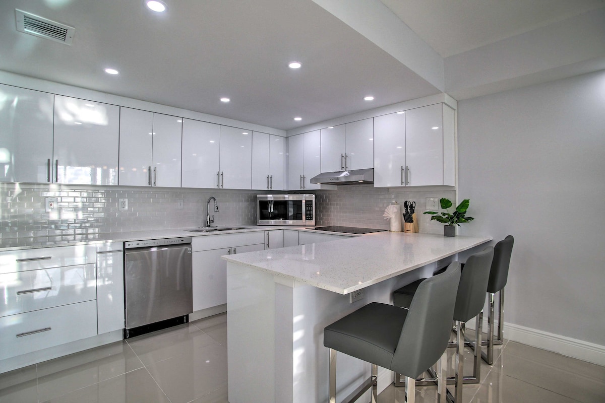 A modern kitchen is presented with sleek white cabinetry and silver appliances. A spacious peninsula counter offers seating for four. The glossy tile flooring reflects ambient light from the recessed lighting above, enhancing the clean and contemporary design.