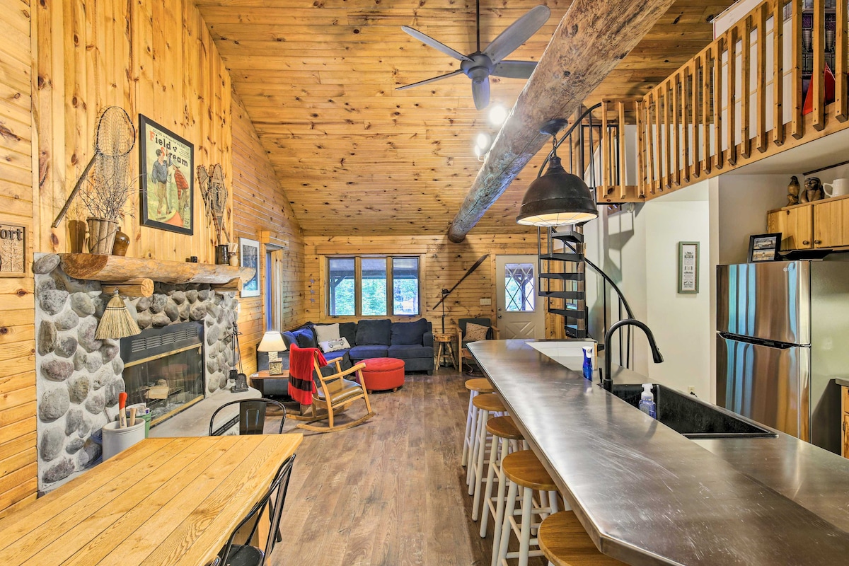 A rustic cabin interior features wooden walls and a high ceiling with exposed beams. The space includes a stone fireplace, a comfortable seating area, and a modern kitchen with stainless steel appliances. A spiral staircase leads to a loft level, enhancing the open layout.