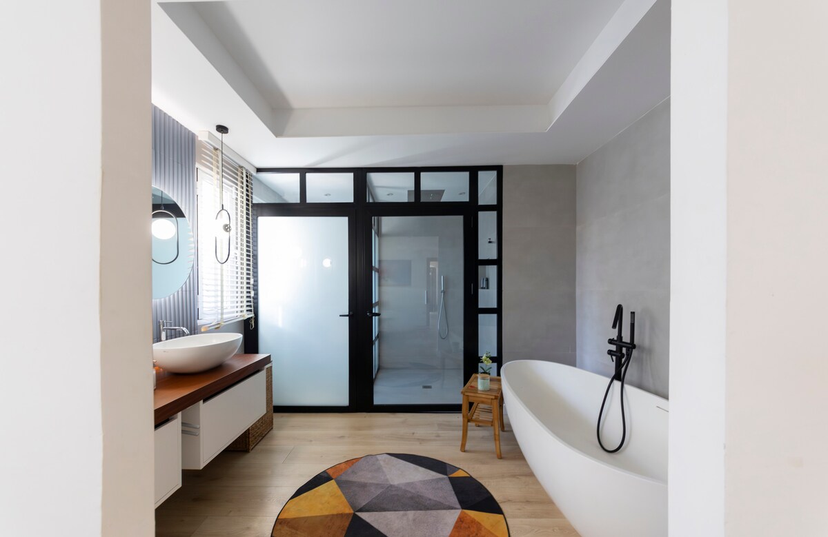 A spacious bathroom features a freestanding bathtub alongside a modern glass-enclosed shower. A round area rug with geometric patterns enhances the floor, and dual sinks are positioned on a wooden vanity. Natural light filters through the window blinds, adding brightness to the space.