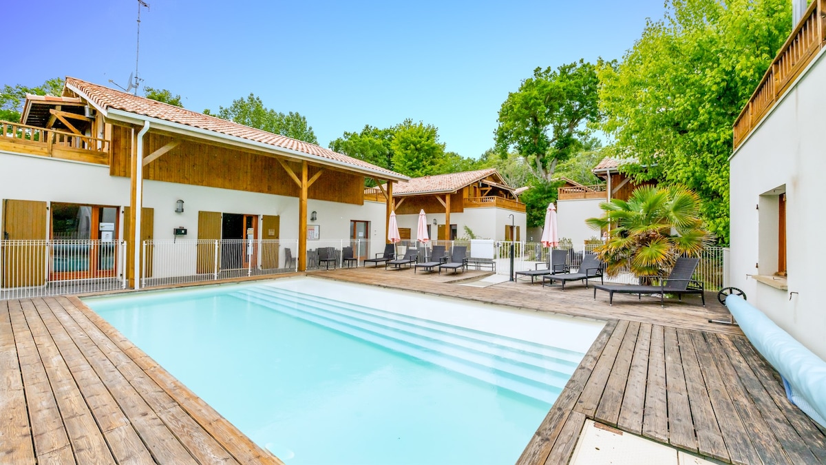 A seasonal outdoor pool is surrounded by wooden decking and sun loungers. Lush greenery provides a natural backdrop, while white-painted buildings with wooden accents are visible in the distance. The pool area is designed for relaxation and enjoyment during warm weather.