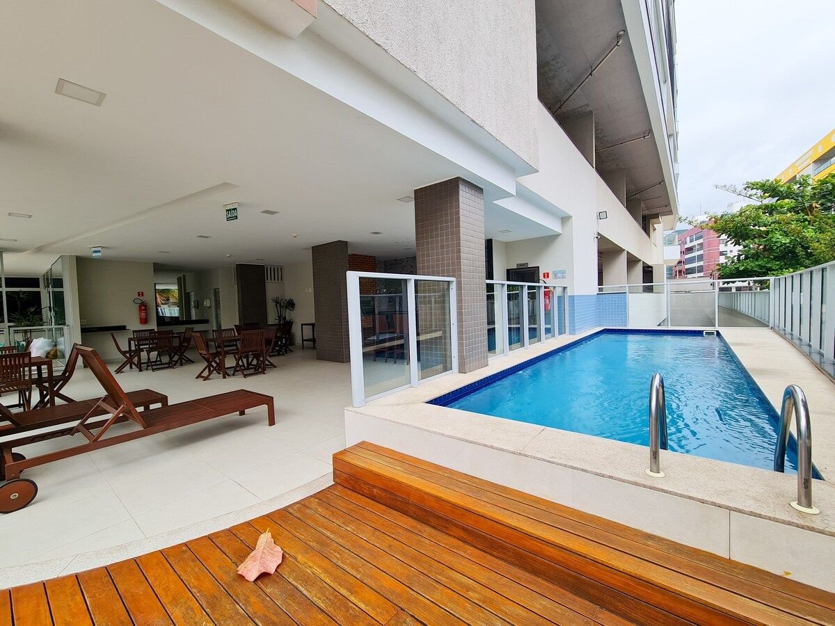 An outdoor area showcases a swimming pool bordered by a tiled deck. Wooden lounge chairs provide seating options beside the pool, while tables and chairs are arranged under a covered space. The setting features a mix of concrete and wood elements, contributing to a functional layout.