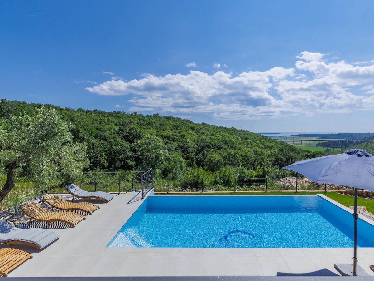 An outdoor area features a rectangular swimming pool with clear blue water, surrounded by wooden deck chairs. Lush greenery spans the landscape in the background, along with a distant view of the sea under a bright blue sky with few clouds.