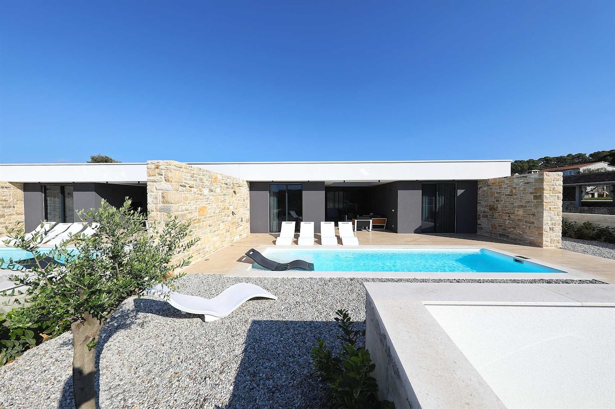 Two identical villas are viewed from the pool area, showcasing modern architecture. A serene pool with lounge chairs is framed by stone walls and a gravel landscape. Clear skies and sunlight create a bright and open atmosphere around the private pools.