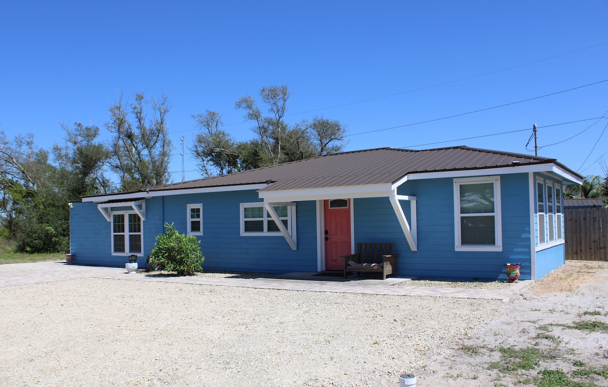 A single-story blue house features a welcoming front porch with a simple bench and a bright door. The property is surrounded by light gravel, and trees provide a natural backdrop. Large windows allow for ample natural light, showcasing the home's spacious design.