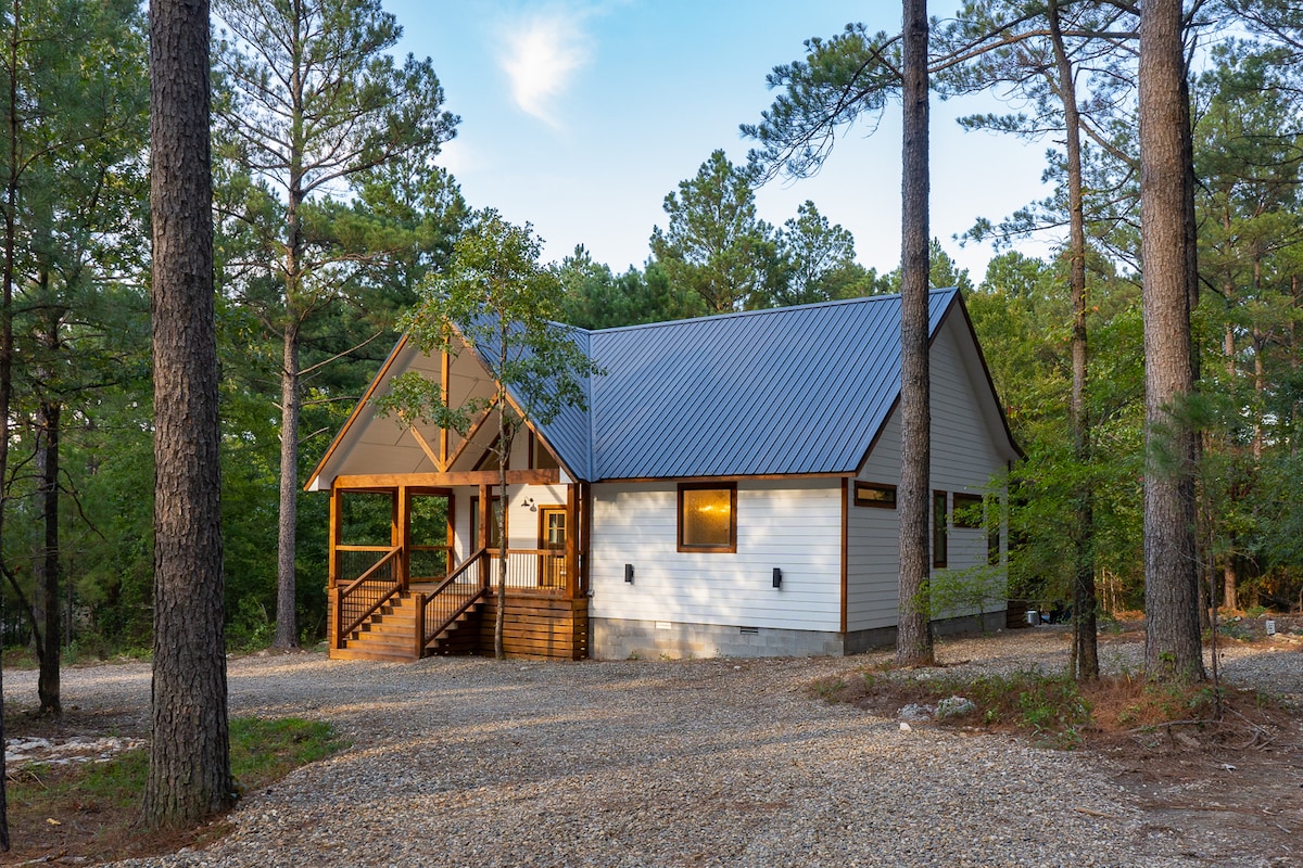 A serene cabin retreat is nestled among towering pine trees, showcasing a welcoming entrance with steps leading up to a covered porch. The exterior features a combination of wooden and white paneling, complemented by a metallic roof reflecting natural surroundings.
