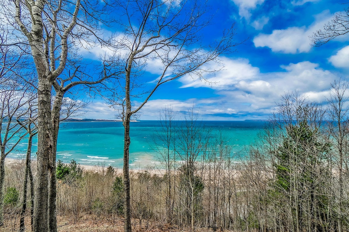 Views of Sleeping Bear Bay and Lake Michigan are captured from a hillside perspective. The water exhibits varying shades of blue, while the shoreline is visible in the distance. Leafless trees and scattered vegetation are present in the foreground, framing the expansive landscape.