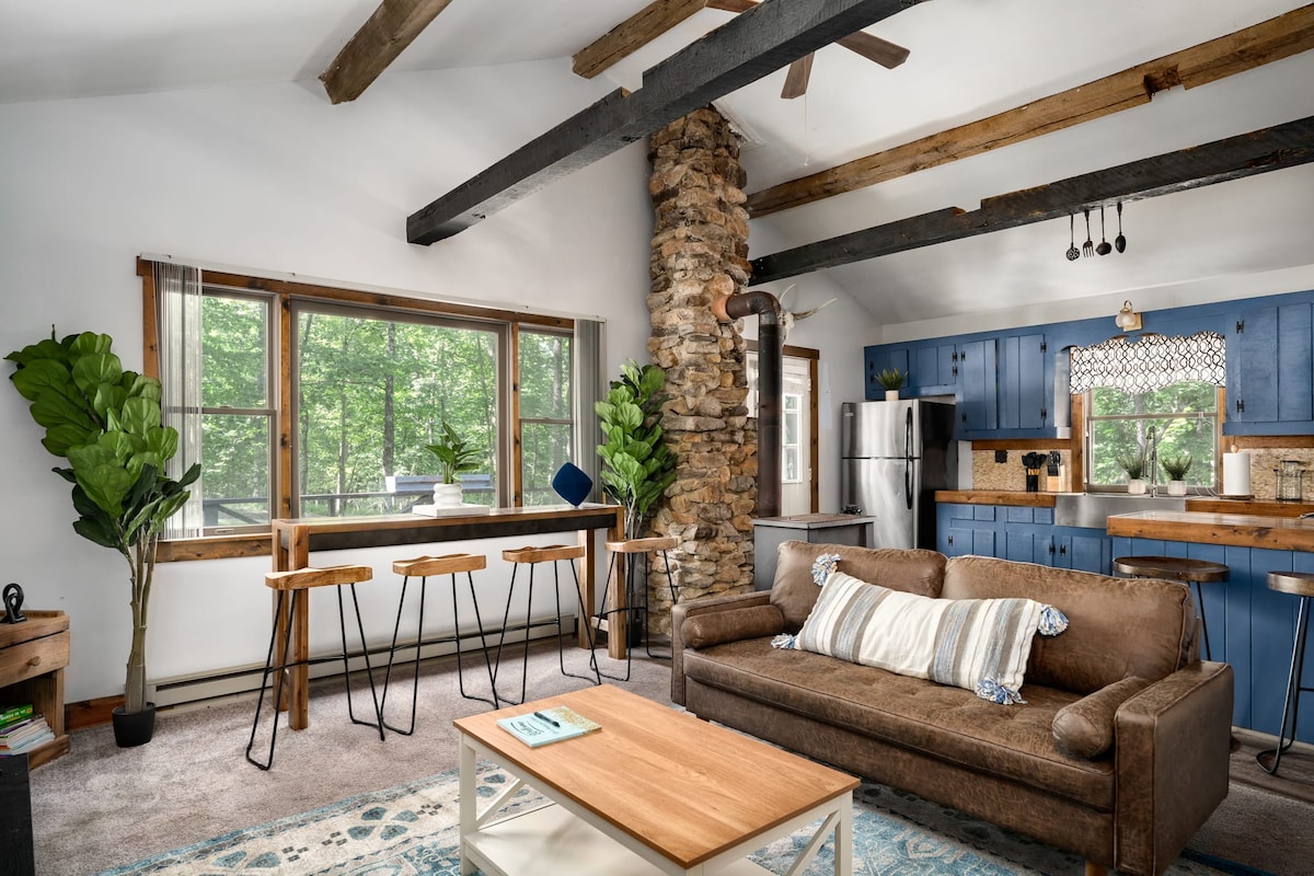 A cozy living area features a brown sofa with blue and white accents, complemented by a light wooden coffee table. Natural light filters through large windows, illuminating the adjacent kitchen with blue cabinetry and stainless steel appliances. A tall houseplant enhances the welcoming atmosphere.
