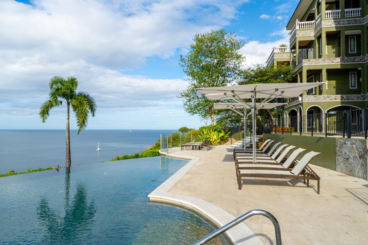 An infinity salt-water pool extends into the ocean view, surrounded by lounge chairs under a large canopy. Lush greenery and distant sailboats can be seen in the calm blue water, complemented by a clear sky.