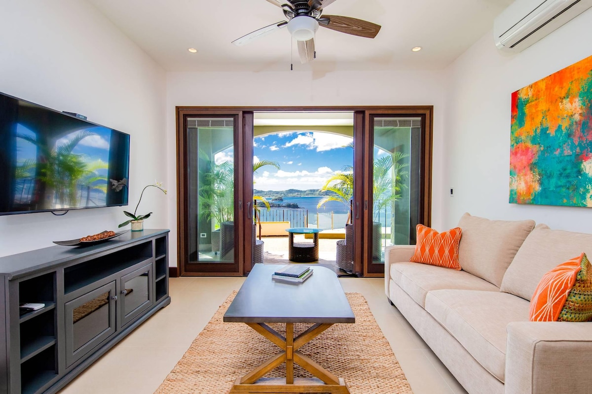 The living area features a comfortable sofa adorned with colorful cushions, a coffee table, and a flat-screen television. Large glass doors open to a stunning ocean view, while bright artwork adds a touch of vibrancy to the neutral décor.