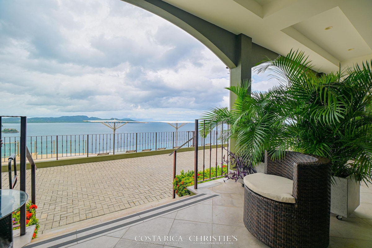 An outdoor terrace features wicker seating, surrounded by lush green plants. A panoramic ocean view is visible beyond the railing, with gentle waves lapping at the shore. The sky above is partly cloudy, adding a serene atmosphere to the coastal landscape.