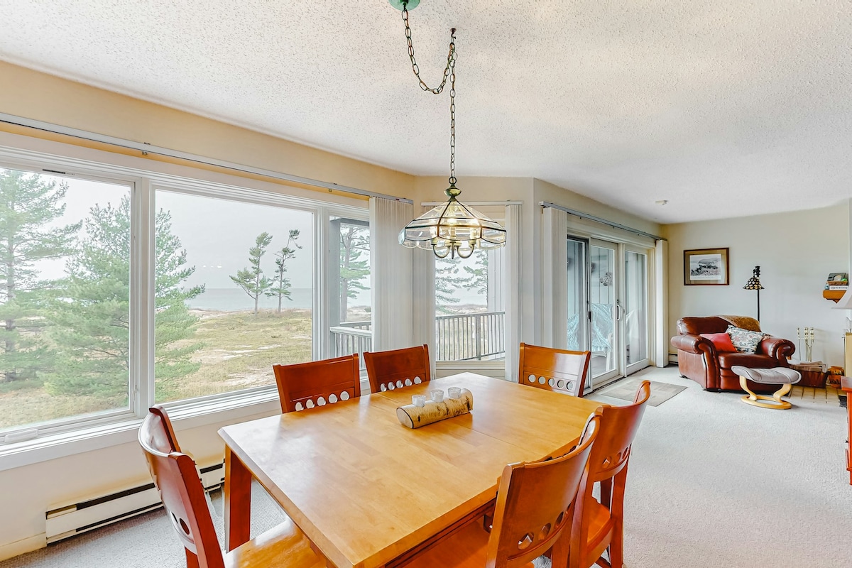 A dining area is furnished with a wooden table surrounded by six chairs. Large windows allow natural light to fill the space, offering views of the outdoor scenery. A comfortable seating area with a leather sofa and side table is positioned nearby, contributing to the open layout.