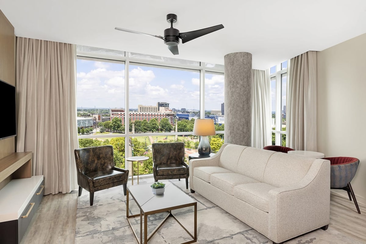 An inviting living area is captured with large windows offering expansive views of downtown Atlanta. A light-toned sofa, two accent chairs, and a coffee table are arranged on a soft area rug, while a ceiling fan adds a modern touch to the bright space.