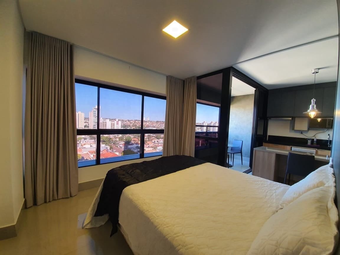 A spacious bedroom features a large window offering a city view. A comfortable queen-size bed is positioned in the center, with light-colored bedding. Dark curtains frame the window, while a kitchenette is visible in the background. Neutral tones create a calm ambiance.