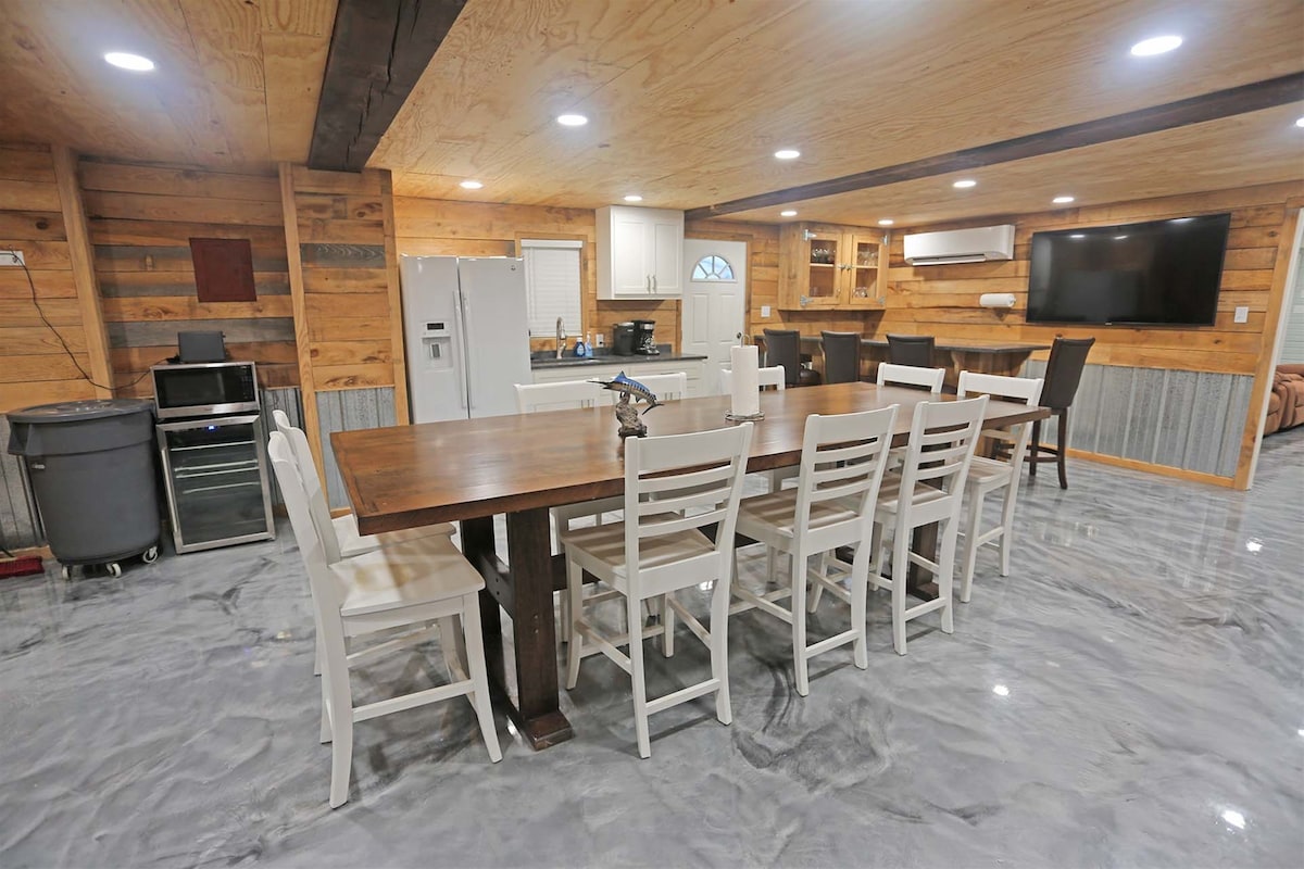A spacious dining area is furnished with a large wooden table surrounded by white chairs. The kitchen, partially visible, features modern appliances, including a refrigerator and a stovetop. Soft lighting enhances the wooden accents throughout the room, providing a warm atmosphere.