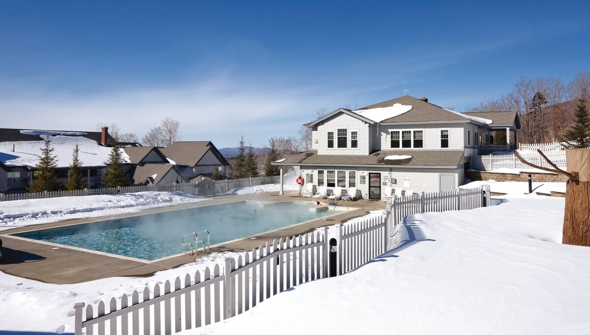 An outdoor swimming pool is surrounded by a white picket fence, with snow covering the ground nearby. The building adjacent to the pool features multiple windows and a covered porch, providing a scenic view of the surrounding area.