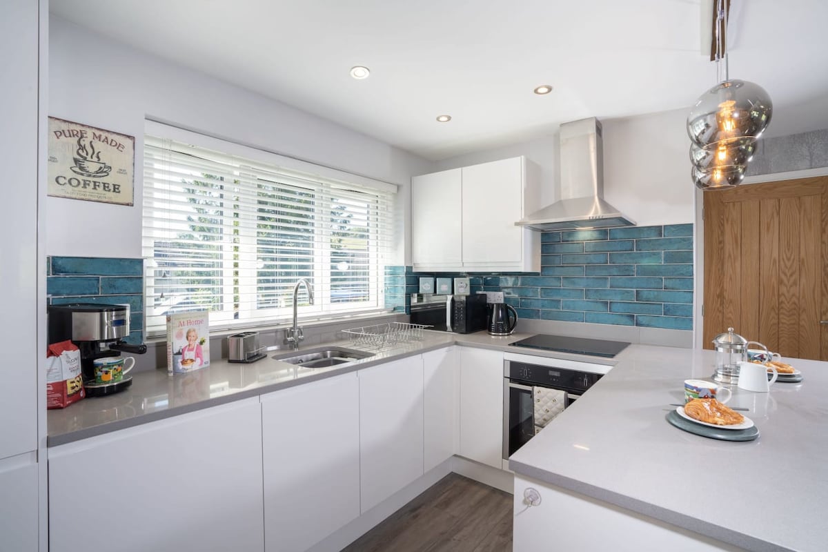 A modern kitchen with sleek white cabinetry and a contemporary design features blue tiled backsplash and stainless steel appliances. Natural light floods the space through a window with white blinds. A breakfast bar is set with plates and drinks, inviting casual dining.